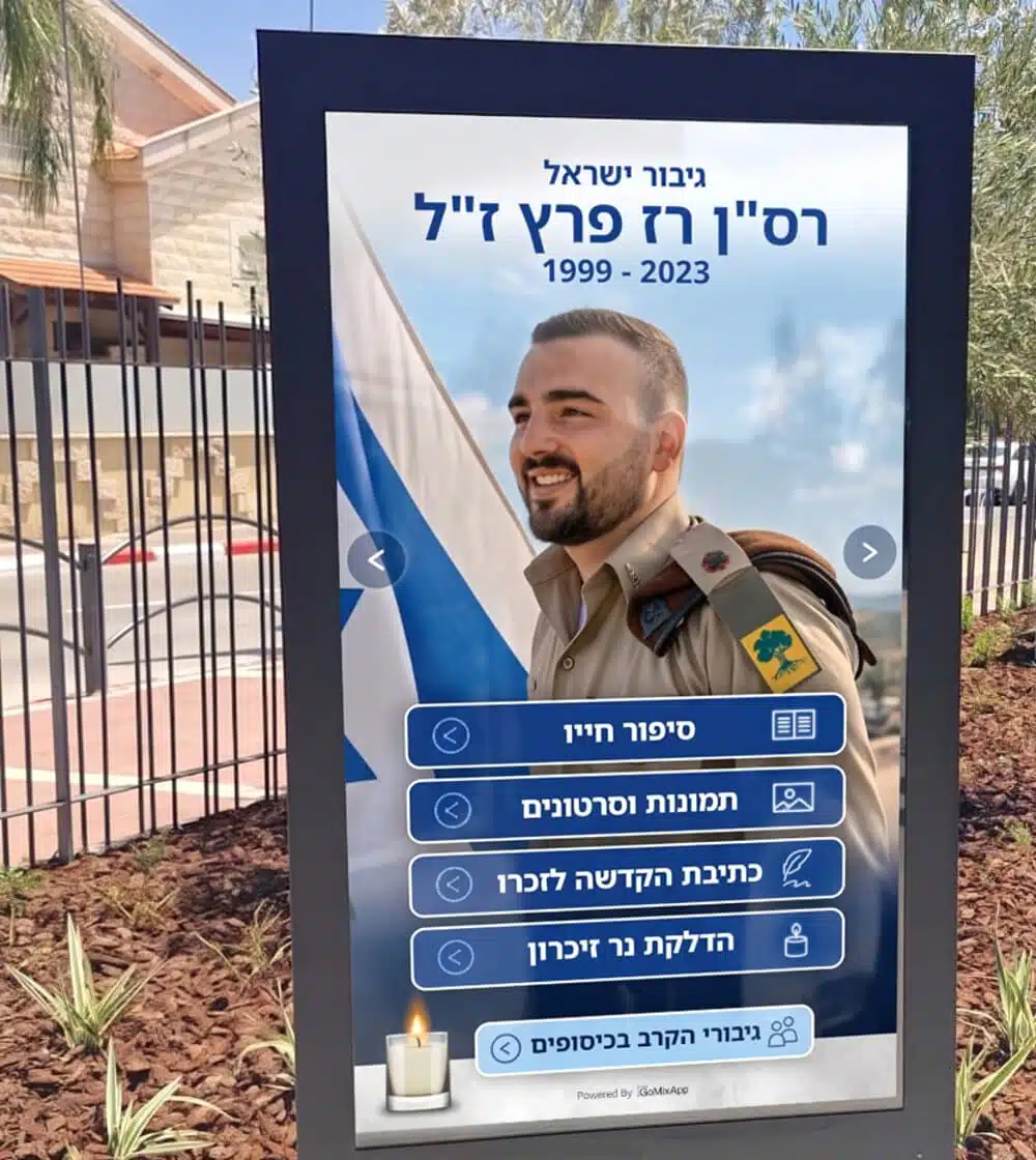 עמדת זיכרון אינטראקטיבית מותקנת באתר הנצחה, מסך מגע חיצוני לצד דגלי ישראל